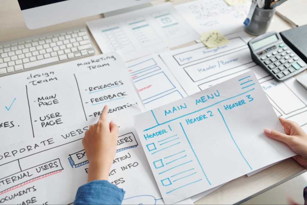 A web designer sketching a website wireframe on paper, mapping out the header, main menu, and user journey.