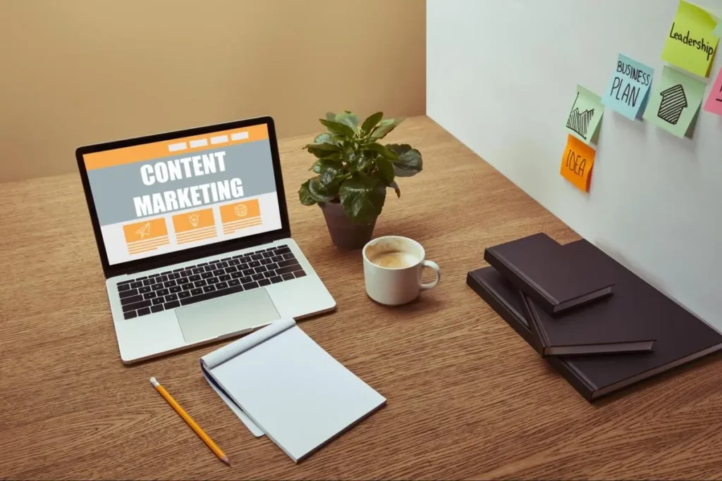 Laptop on a wooden desk displaying a content marketing webpage, next to a notebook, coffee cup and stack of books